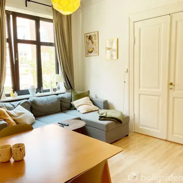 Sofa med puder står i hjørne ved vindue; gardiner hænger ned. Stuebord har to krus; vægmaleri og dør til højre. Lys træbund fuldender rummet.