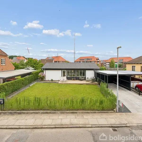 En moderne, lavbundet bolig med fladt tag står bag en velplejet græsplæne. En rød bil holder parkeret i carporten til højre. Klar himmel med få skyer.
