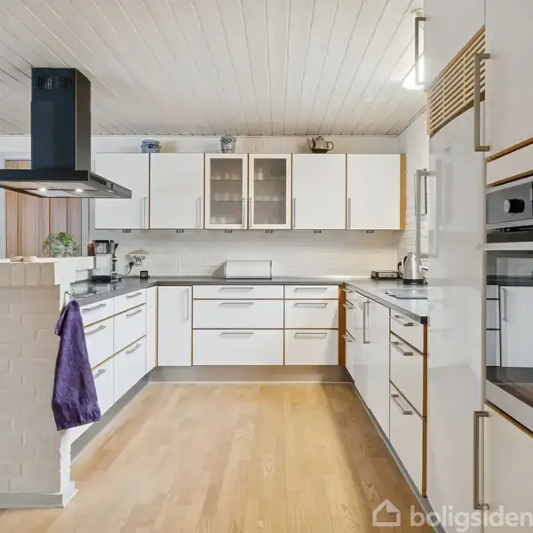 Et moderne køkken med hvide skabe og sorte bordplader. En indbygningsovn er integreret i skabsvæggen. En lilla klud hænger fra en hvid murstensskillevæg i et lyst rum.