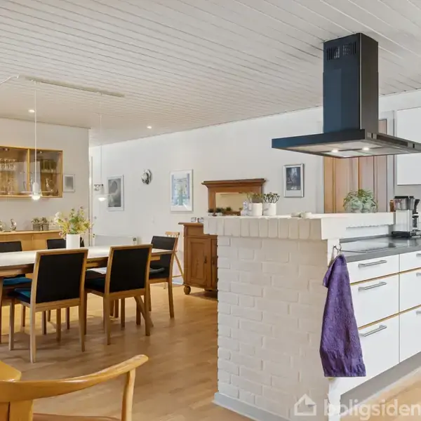 Et moderne køkken med hvide skabe og en sort emhætte, hvor en lilla håndklæde hænger, ligger i åben forbindelse med en spisestue med et træbord og sorte stole.