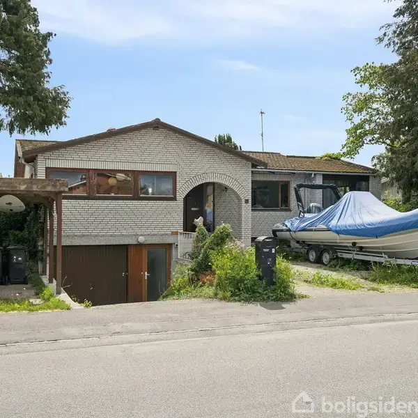 Et hvidt murstenshus står med en garage nedenunder. En båd er placeret på en trailer i indkørslen. Omkring er der træer og buske.