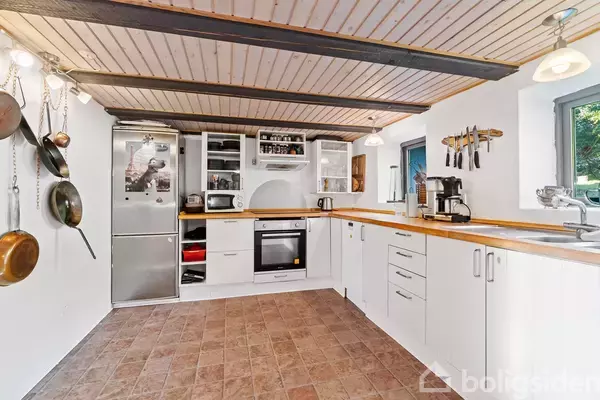 Et lyst køkken med hvide skabe, træbordplader og en indbygget ovn; køleskab til venstre; pander hænger ved siden af. Flisegulv og træbeklædt loft med lamper. En kaffemaskine står ved vinduet.