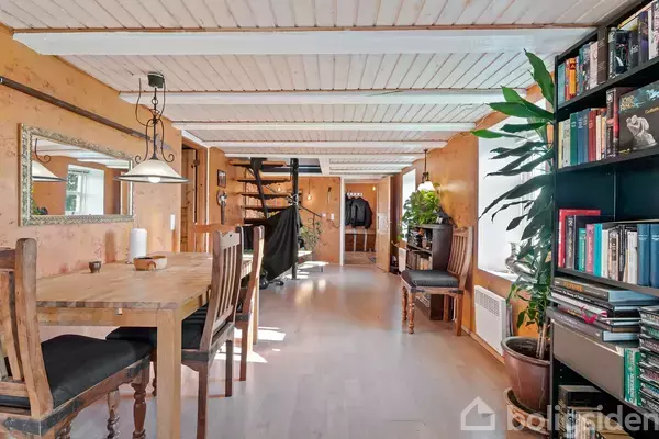 Et træbord med stole står i en lang korridor. En spiraltrappe er synlig i baggrunden, omgivet af planter og bogreoler. Loftslamper lyser rummet op.