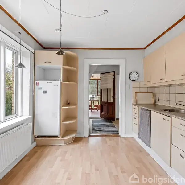 Køkken med lyst trægulv og beige skabe langs væggen. Et køleskab står ved siden af et stort vindue. En dør i baggrunden fører til et andet rum.