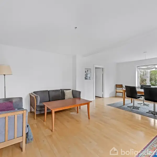 En lys stue med træmøbler står stille på et lyst trægulv. Sofa og bord til venstre, spisebord med stole til højre ved vindue. Lampe og tæppe skaber hygge.