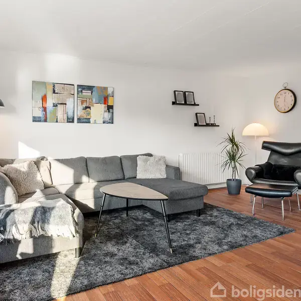 Grå sofa står i en stue, suppleret af en lænestol og moderne kunst på væggen. Stueetagen har trægulve, et rundt vægur og en sort gulvlampe ved siden af sofaen.