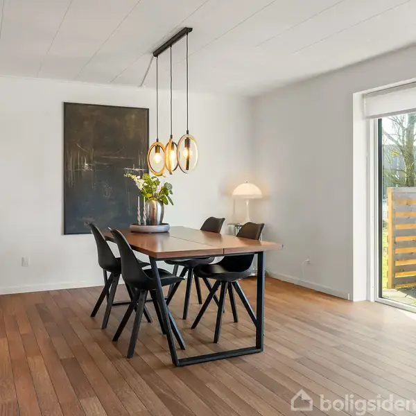 Spisebord med fire stole står centralt under tre hængende lamper. Et stort maleri hænger på væggen. Ved siden af er en plante og en lampe. Lys strømmer ind gennem terrassedøre.