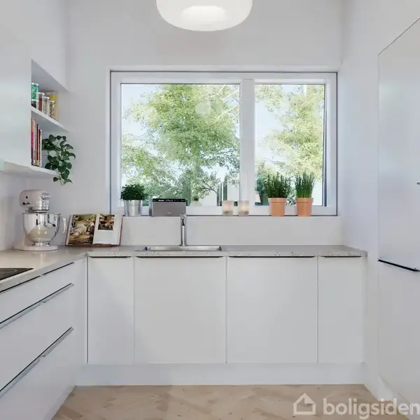 Køkkenet har hvide skabe og en bordplade med køkkenredskaber og en brødrister. Der er en stor vindue med udsigt til træer, og ovne i væggen til højre.