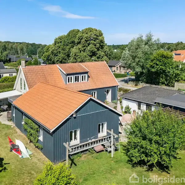 Huset har en rød tag og blå vægge, omgivet af en grøn have med træer og buske, under en blå himmel. Flere andre huse ses i nabolaget.