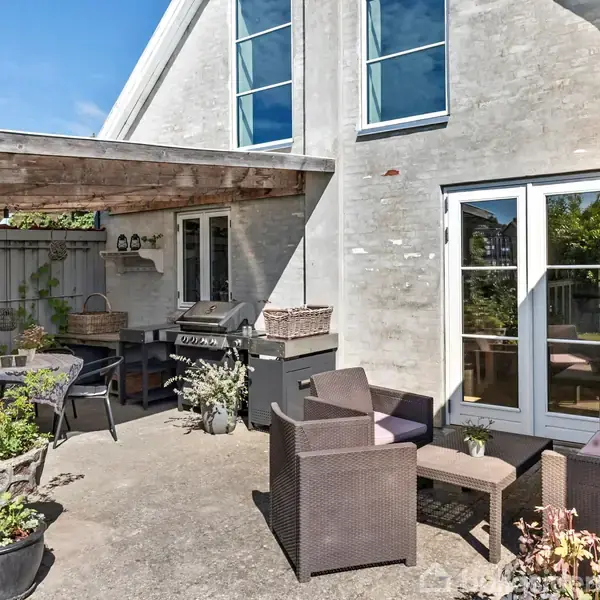 Terrasse med møbler af mørk flet står under en pergola ved et moderne hus. En grill står nær et bord med stole, omringet af planter i krukker.