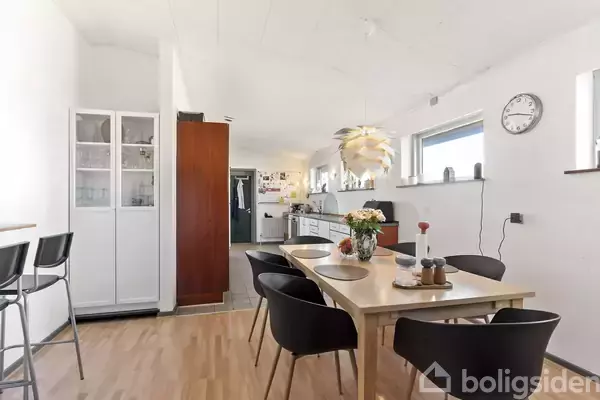 Et spisebord er dækket med tallerkener og en vase med blomster, omgivet af sorte stole. Køkken med skabe og barstole i baggrunden. Ur på væggen viser klokken 10:08.