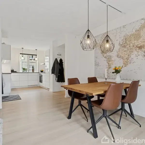 Spisebord med fire stole står under to hængelamper. I baggrunden ses et kort på væggen og åbent køkken. Lys indretning med sofa og tæppe til venstre.