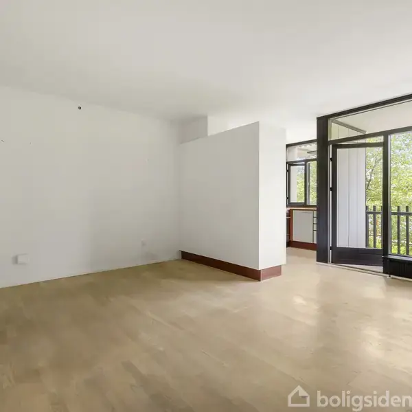 Balkon med mørkt rækværk, vender ud mod grønne træer og en blå himmel, set fra en åben dør. Der er et betongulv og hvid ydervæg til venstre.