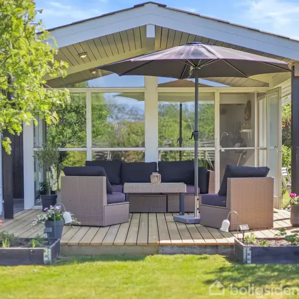 Terrassemøbler med sofaer og et bord står under en stor parasol. De er placeret foran et moderne, åbent havehus med glasvægge, omgivet af frodig grøn beplantning.
