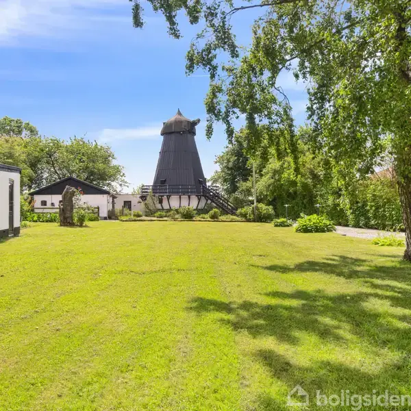 En sort mølle står i en grøn have omgivet af træer og buske. Ved siden af ses et hvidt hus. Skyfri himmel og sollys oplyser scenen.