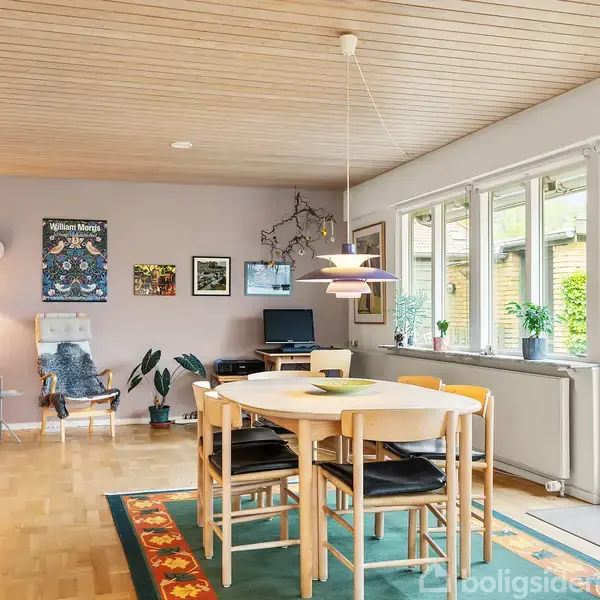 Rundt spisebord med stole står på et farverigt tæppe i en lys stue med trægulv og loft. Store vinduer til højre bringer naturligt lys ind. Vægkunst og reoler pryder rummet.