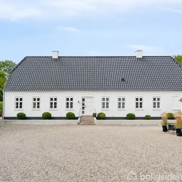 Et hvidt, traditionsrigt landhus med sort tegltag står foran en gårdsplads med grus. Flere buske er plantet langs husets facade. Bygningen er omkranset af bygninger med stråtag.