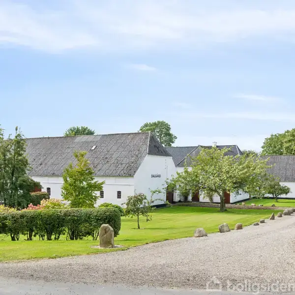Hvidt, stråtækt hus ligger i en grøn have med buske og træer. En grusvej fører til hovedindgangen, omgivet af sten og velplejet græsplæne.