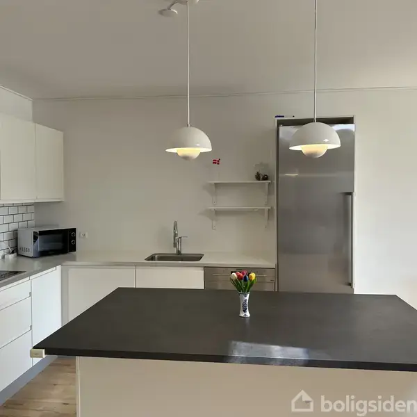 Køkken med sort køkkenø og hvidt interiør, to hængende lamper og vase med tulipaner, moderne ovn, mikroovn, stål køleskab, hvid flisevæg og trægulv.