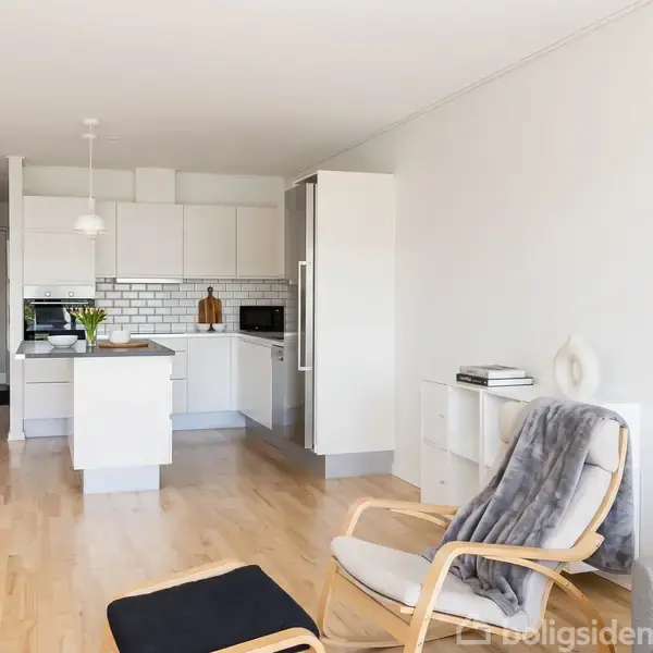 Et lyst køkken-alrum med trægulv indeholder et hvidt køkken med en ø, moderne apparater og små fliser. En lænestol med tæppe og fodskammel står i opholdsområdet.