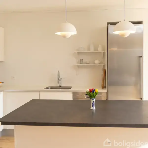 Køkken med hvidt interiør, en sort bordplade med en vase med tulipaner, og en køkkenvask ved siden af et stort køleskab i rustfrit stål. To lamper hænger over bordet.