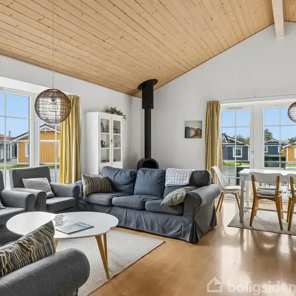 Stue med grå sofaer og et rundt sofabord, pejs i hjørnet. Ved siden af er spisebord med stole. Store vinduer slipper lys ind. Husene udenfor ses.