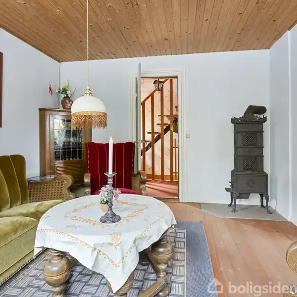 En stue med en grøn sofa står foran et træbor med en hvid dug. Til højre er en gammel komfur, og baggrunden viser en trappe. Loftet er af træ.