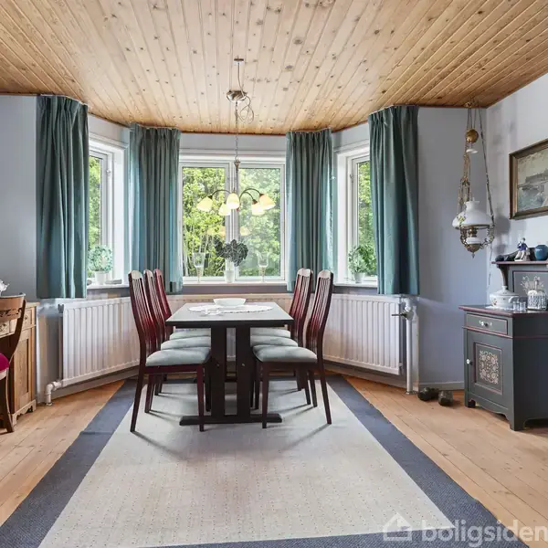 Spisebord med stole er placeret nær vinduer med grønne gardiner. Rummet har træloft og en blå væg. En åbning fører til en stue med sofaer og dekorationer.