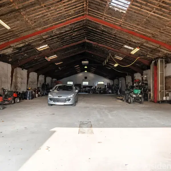 Et stort lager indeholder en bil i midten og forskellige maskiner langs siderne, opbevaret under et højt træloft med synlige bjælker og spredt belysning, i et industrielt miljø.