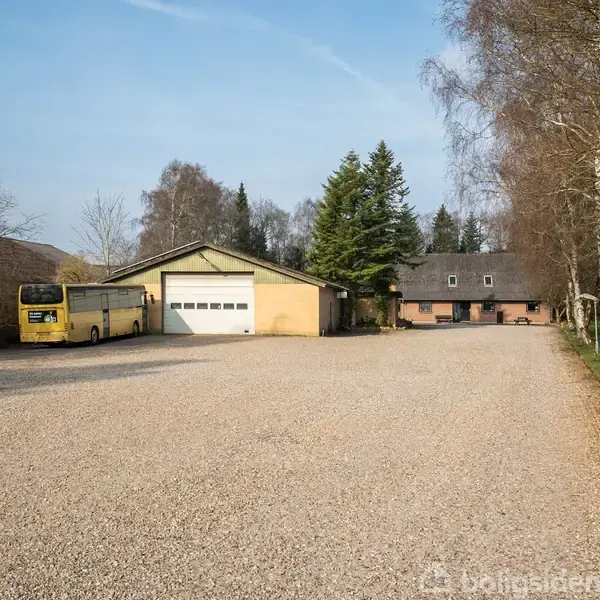 En gul bus holder parkeret ved siden af en stor garage med hvide døre. I baggrunden ses en bygning med skråtag omgivet af træer og grusplads.