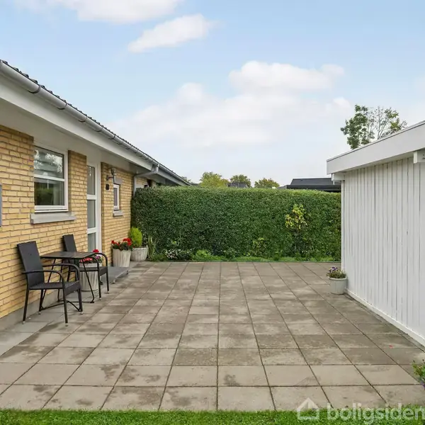 Hus med gul teglvæg og fladt tag, står ved siden af en belagt terrasse. To sorte stole ved bordet. Høj hæk i baggrunden og blomsterbede ved siden af.
