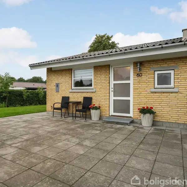 Gulstenshus med to stole og et bord foran, står stille i en have. Husnummer 60 er ved hoveddøren. Fliser dækker terrassen, omgivet af græsplæne og buske.