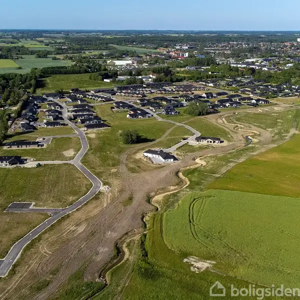 Nyudviklet boligområde med veje, huse under opførelse, omgivet af marker og et fjerntliggende byområde. Grusveje snor sig gennem området, der grænser op til grønne marker.