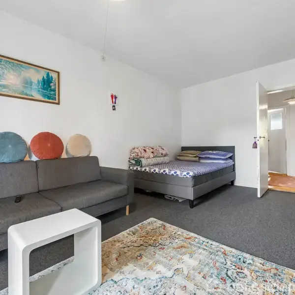 Sofa med farverige puder står i stue ved siden af en seng med blomstrede sengetøj. Et lille bord står på et mønstret tæppe. En åben dør fører til en gang.