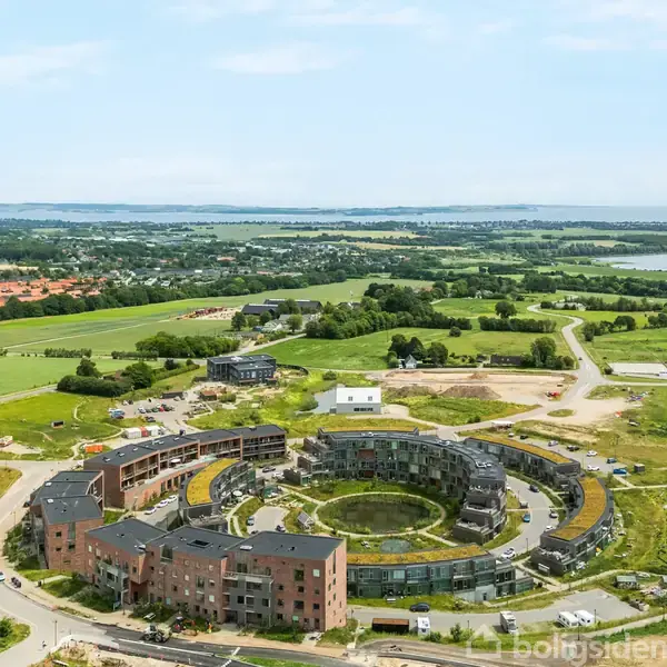 Rund bygning med grøn tagflade omgiver en central gård, placeret i landligt område med marker og spredt beboelse i baggrunden, under en klar blå himmel.