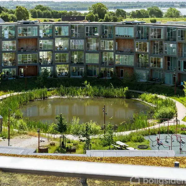 Et moderne, cirkulært lejlighedskompleks omslutter en gård med en dam og grønne områder. Der er et shelter og en legeplads i forgrunden, omgivet af stier og planteliv.