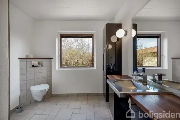 Et moderne badeværelse har hvide fliser og en væghængt toiletkumme. En sort skabsmodul med spejl og vask er oplyst af væglamper. Store vinduer giver udsigt til træer udenfor.