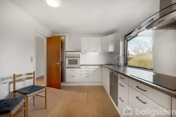 Et lyst, moderne køkken med hvide skabe og en indbygget ovn; store vinduer som giver udsigt over træer. To stole står langs væggen, og gulvet er i træ.