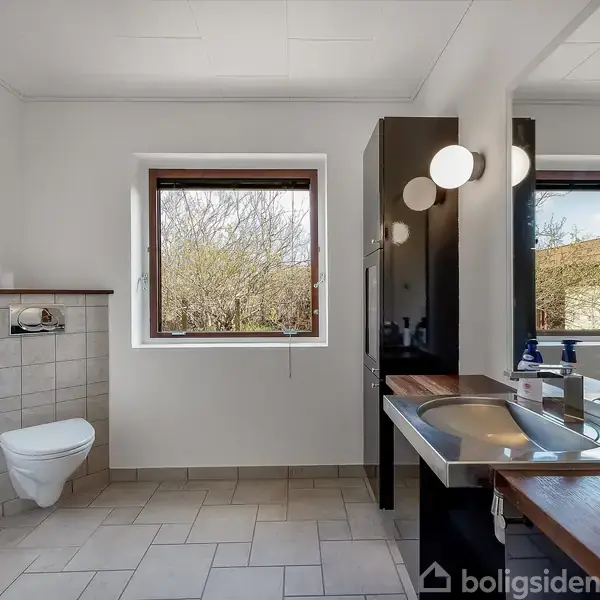 Toilet monteret på væg, ved siden af et vindue med udsigt til buske. Håndvask med spejl og hylde, væglamper over, i et lyst badeværelse.