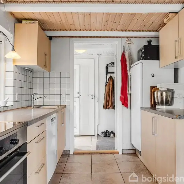 Et køkken med lyse skabe, en indbygget ovn og keramiske fliser på væggen. En mixer og krukker står på bordet. En åbning fører til en entré med jakker.