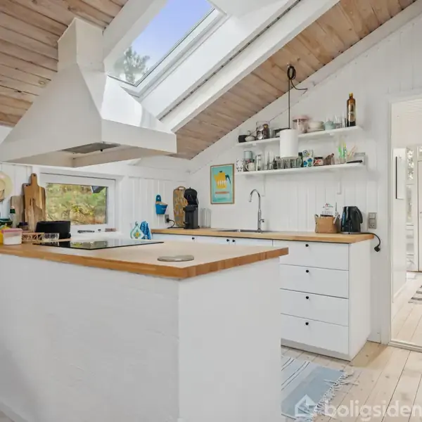 Køkken med hvidmalede skabe og træbordplader ligger i en lys, skrånende loftsrum. Ovenlysvinduer lukker dagslys ind. I baggrunden ses en åben dør til en veranda.