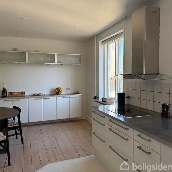 En moderne køkken med hvide skabe og sølvhåndtag. Bordpladen har en indbygget kogeplade, og der er en emhætte ovenfor. Lyse trægulve og et firkantet spisebord med stole i hjørnet.
