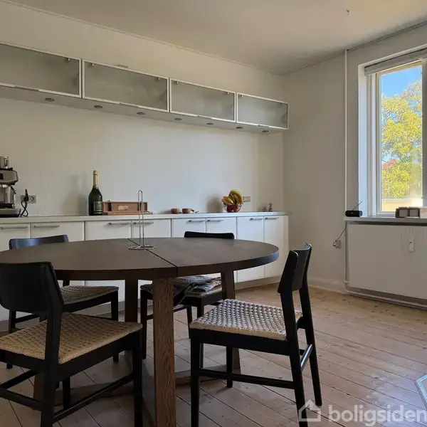 Et rundt spisebord med fire stole står i et lyst køkken. På bordet er der en kageopsats. Bagved ses en kaffemaskine, vinflaske og dekorationer på en skænk.