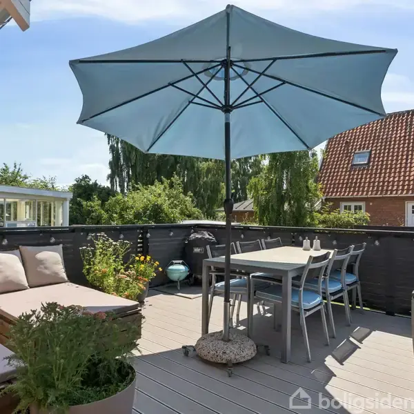 Terrasse med bord og stole under en stor, blå parasol. To liggestole med puder og planter ses i et afslappende udemiljø oppe på taget med udsigt til omkringliggende huse.
