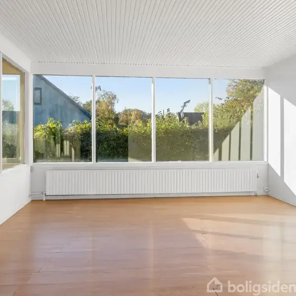 Et lyst, tomt rum med trægulv og store vinduer, der vender mod en have med buskads. Hvide vægge og et radiatorpanel under vinduerne.