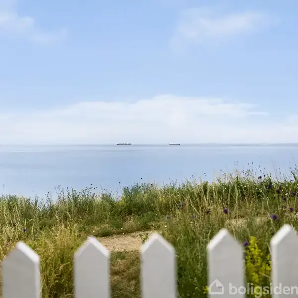 Hvidt stakit står i forgrunden, hvor det adskiller en sti fra en blomstrende mark og rolige havudsigt i baggrunden under en lyseblå himmel.
