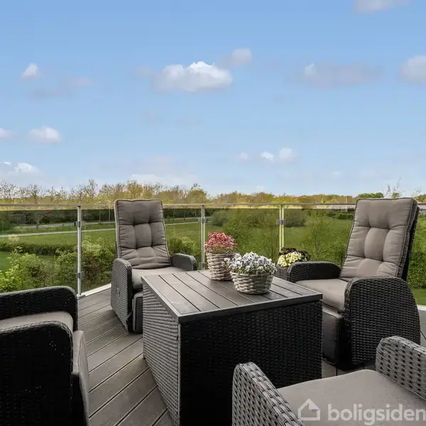 Terrassemøbler i rotting står rundt et bord med blomster, placeret på en terrasse med glasrækværk, der giver udsigt over en grøn have og en klar blå himmel.