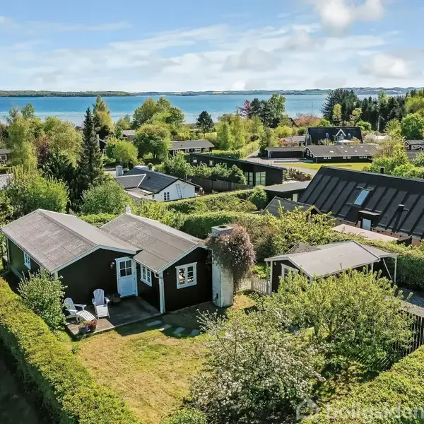Lille hus omgivet af hække og træer, med terrassebord foran. Huset ligger i et grønt område med flere huse i nærheden. I baggrunden ses fjorden og skovklædte områder.