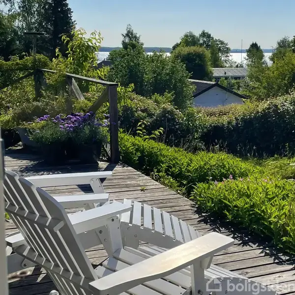 Hvide liggestole står på en træterrasse omgivet af grønne planter og blomsterkrukker med lilla blomster. I baggrunden ses et hus og træer med vand i horisonten.