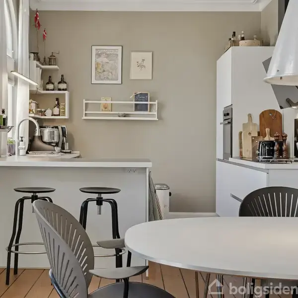 Et lyst køkken med moderne møbler; to barstole ved en hvid bardisk, køleskab og indbygget ovn til højre. Hylder over vasken indeholder dekorative genstande. Ovalt spisebord foran.